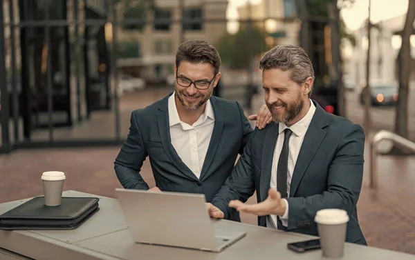 Neşeli iş adamları bilgisayarla çevrimiçi ticaret yapıyor. İnternet üzerinden ticaret yapan iş adamlarının fotoğrafı. İki işadamı çevrimiçi ticaret yapıyor. İş adamları açık havada ticaret yapıyor.