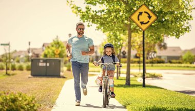 Baba ve oğul bisiklet macerasında. Baba ve oğul ikilisi resimli manzarada pedal çeviriyor. Destekleyici baba oğluna ilk bisiklet sürüşünde rehberlik ediyor. Baba oğul bisiklet gezintisinin keyfini çıkarıyorlar. Aile Vakfı.