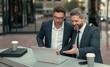 Gülümseyen iş adamlarının internetten alışveriş yaptığı fotoğraf. İki işadamı çevrimiçi ticaret yapıyor. İş adamları açık havada ticaret yapıyor. İşadamları bilgisayarla online ticaret yapıyor.