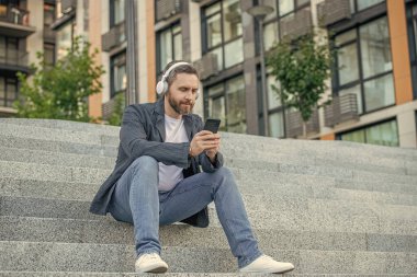 Adam müzik listesini seçiyor. Açık havada oturuyor. Adam sokakta müzik listesi seçiyor. Dışarıda müzik listesi seçen bir adam var. Bir adamın fotoğrafı telefonda müzik çalma listesini seçin.