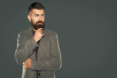 Düşünceli, sakallı, olgun bir adam gri arka planda rahat giyinir..