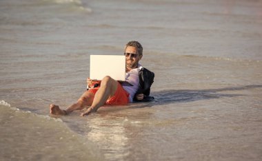 Uzaktan kumanda çalışıyor. İş adamı plajda rahatlar. Yazın dizüstü bilgisayarı olan takım elbiseli bir iş adamı. İş adamı yazın denizde iş yapmayı hayal eder. seyahat ve rahatlama. Kumsalda dinlenen bir iş adamı..