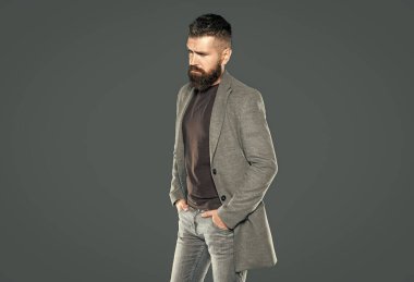 Hırslı, sakallı, olgun bir adam gri arka planda rahat giyinir..