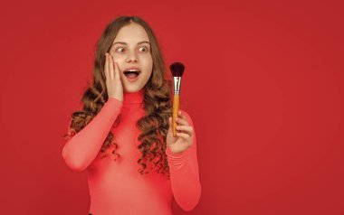 Şok olmuş kıvırcık saçlı kız makyaj için pudra fırçası tutuyor..