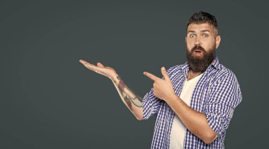 Şaşırmış sakallı adam kareli gömlek giyer. Gri arkaplanda kopya alanı olan ürünü sunar..