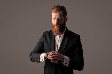 Saati ve smokini kontrol eden dakik bir adam. Smokin modası. Smokinli dakik sakallı adam gri arka planda izole edilmiş saati kontrol ediyor. Smokin giyerken çekilen dakik adam stüdyosu kontrol saati.