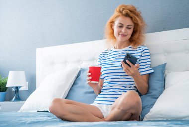 Telefonda internette kahve sörfü yapan neşeli bir kız. Kız evde telefonuyla internette sörf yapıyor. Kızın evinde internet sörfü.