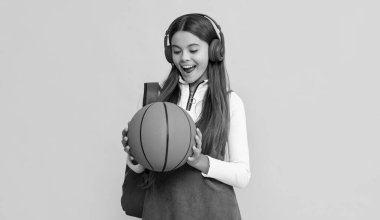amazed kid in headphones with school backpack and basketball ball on yellow background.