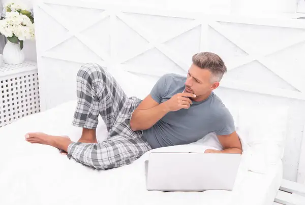 pondering man freelancer at home in bed. man freelancer at home in bedroom. photo of man freelancer at home with laptop. man freelancer at home.