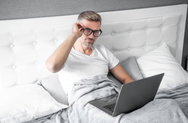 Gözlüklü olgun bir adam laptopla yatakta çalışıyor. siber pazartesi.