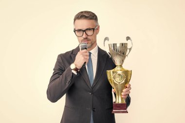 Takım elbiseli olgun bir adam şampiyon kupası ve beyaz arka planda izole edilmiş mikrofon tutuyor..