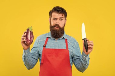 Puzzled man in apron holding chefs knife and eggplant yellow background, chef.