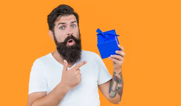 surprised birthday guy holding present box. bearded guy with present box in studio. guy with present isolated on yellow background. present box for guy in shirt.