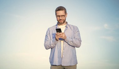Gözlüklü adam akıllı telefondan mesajla sohbet ediyor. Adam telefonda mesajlaşıyordu. Akıllı telefon iletişimi. Adam gökyüzü arka planında sohbet ediyor.