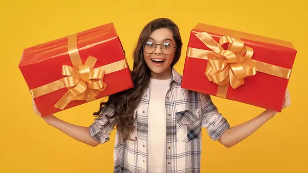 surprised teen kid in glasses with present. studio shot of teen kid hold present. teen kid holding box of present. teen kid with present isolated on yellow background.
