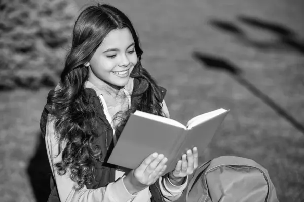 photo of positive school teen girl read in park. school teen girl read outdoor. school teen girl read outside. school teen girl read book.