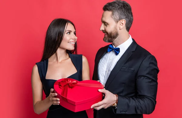 Loving man giving present gift box for Valentines day to surprised woman. Man receiving present. Happy Valentines day. Couple in love isolated on red. Birthday couple with gift. For his sweetheart.