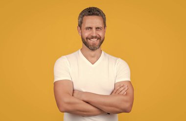 Happy man yellow studio background. Grizzled man smiling in casual white tshirt. Handsome man keeping arms crossed.