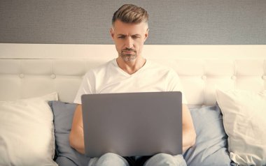 mature freelancer working on laptop in bed.