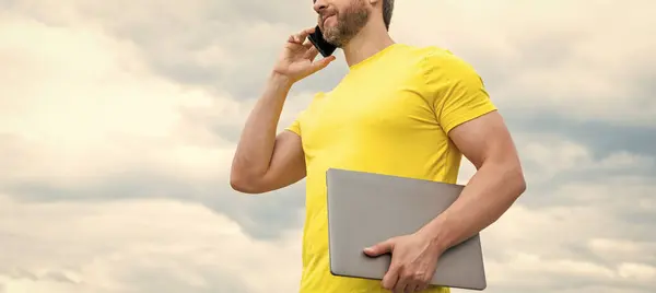 cropped view of man with laptop talking on smartphone on sky background.