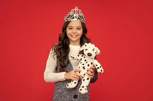 cheerful child in queen crown. princess in tiara. kid hold toy. teen girl wear diadem on red background. happy childhood.
