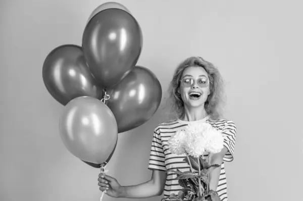 surprised girl with balloons at mothers day. photo of girl with balloons at mothers day. girl with balloons and flowers at mothers day isolated on yellow. girl with balloons at mothers day in studio.