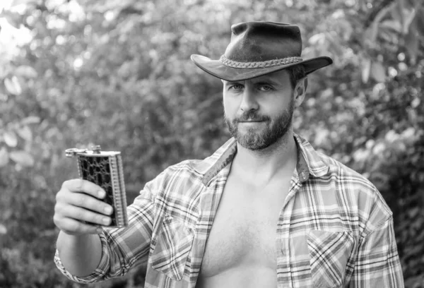 photo of western man offering whiskey flask. western man with whiskey flask. western man with whiskey flask outdoor. western man with whiskey flask wear checkered shirt.