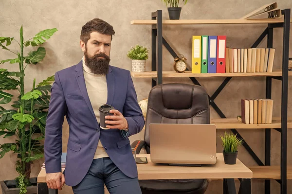 Serious manager in suit drinking tea or coffee sitting on office desk, break.