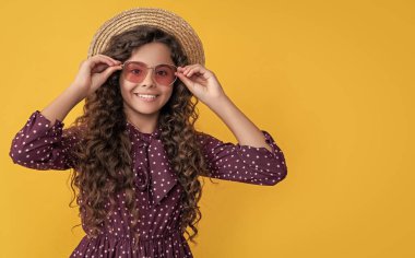 Hasır şapkalı ve güneş gözlüklü mutlu çocuk. Sarı arka planda uzun sarı kıvırcık saçlı..