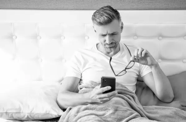 concentrated man in bed read phone wearing eyeglasses.