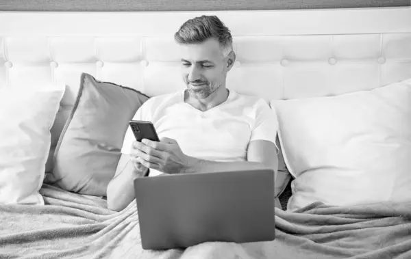 smiling mature man chatting on phone and working on laptop in bed.