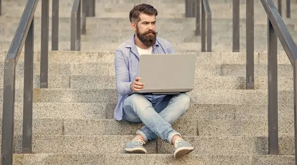 guy has online video pc communication on laptop. guy has online video communication outdoor. guy has online video communication outside. guy has online video communication sitting on stairs.