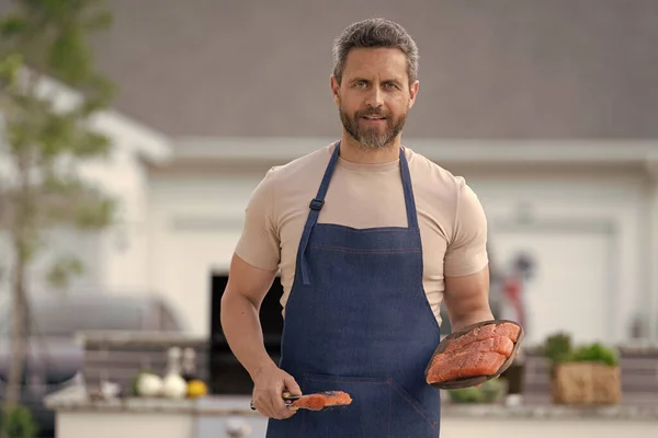 photo of smiling man cook salmon food. man cook salmon on grill. man cook salmon outdoor. man cook salmon in apron.