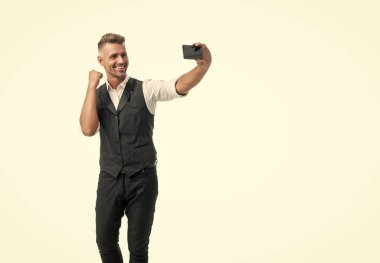 Takım elbiseli mutlu bir çalışan, beyaza izole edilmiş bir cep telefonundan zafer jesti yapıyor..