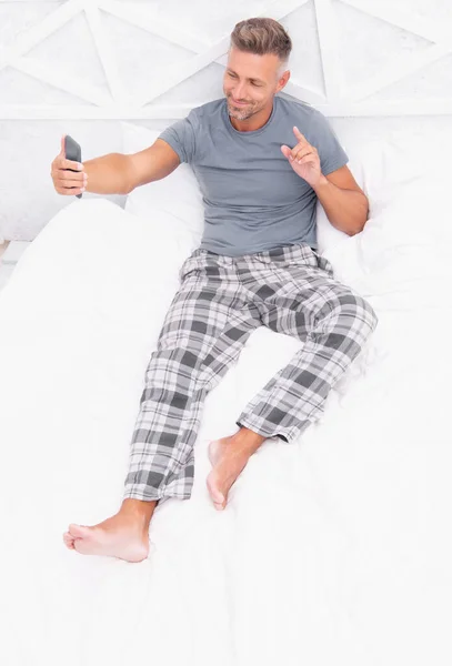 video call man with phone in bedroom, raised finger. video call man with phone wear pajama. video call man with phone in bed. photo of video call man with phone at home.