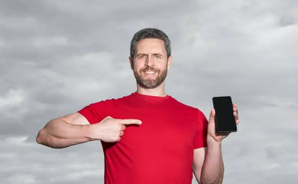 happy man showing phone app on sky background. man showing phone app outdoor. man showing phone app wear red tshirt. photo of man showing phone app.