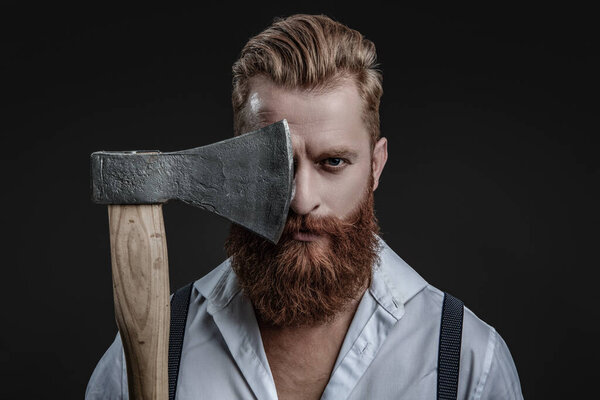 Brutal man in suspenders with ax isolated on black. Mature redhead man with hairstyle. Brutal male fashion style. Male brutality. Vintage brutality guy with axe in retro suspenders. Straight look.