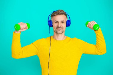 Olgun bir adam mavi üzerine izole edilmiş spor eğitimi yapar. Halterli ve dambılı bir sporcu. Müzikli spor salonu. Spor salonu. Atletik egzersiz için müzik. Güçlü sporcu adam..