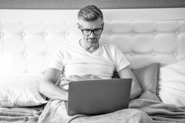 Gözlüklü olgun bir adam laptopla yatakta çalışıyor. dijital göçebe.