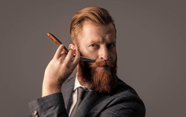 Adamın stüdyoda bıyığı ve sakalı var. Tıraş olmak için bıçağı tutan adam. Sakal tıraşı kavramı. Sakallı adam, gri arka planda sakallı, bıçaklı bıyıklı. Bıçakla bıyık kesen adam..