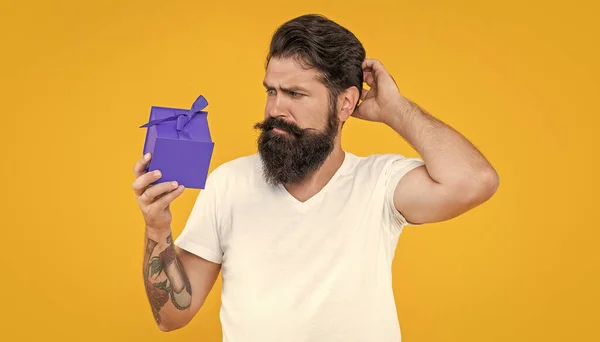 puzzled guy with present isolated on yellow background. present box for guy in shirt. birthday guy holding present box. bearded guy with present box in studio.