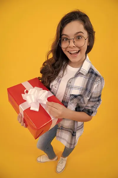 amazed teen kid in glasses with present. studio shot of teen kid hold present. teen kid holding box of present. teen kid with present isolated on yellow background.