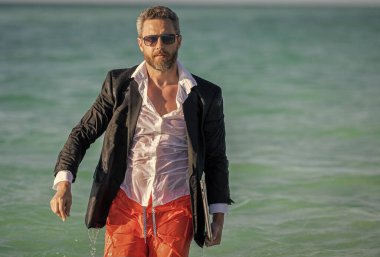 İşletme patronu hafta sonunu sahilde geçiriyor. İş adamı takım elbiseleri sahilde rahatlar. Resmi olarak dizüstü bilgisayarı olan iş adamı tropik plaj yakınlarında giyiniyor. Sıcak iş yazı. Kendini rahatlamaya bırak..