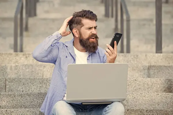 Bearded man reading shocking news on mobile phone. Shocked man looking at mobile phone working on laptop. Mobile internet user. Online news on mobile device.