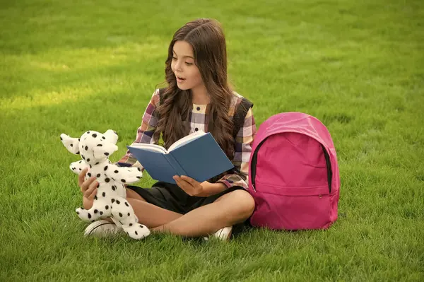Teenage girl reading book to dog toy sitting on grass, school. back to school. pupil at school time.