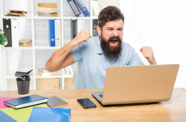 Heyecanlı adam dizüstü bilgisayarda bağırmaktan mutlu. Ofis masasında zafer jesti yapıyor..