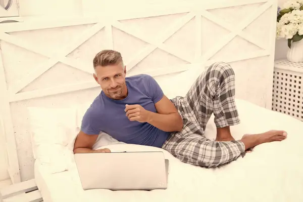 Evde, yatak odasında serbest çalışan neşeli bir adam. Evde dizüstü bilgisayarlı serbest çalışan bir adamın fotoğrafı. Evde serbest çalışan bir adam var. Adam evde, yatakta serbest çalışıyor..