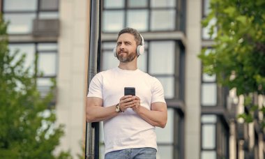 photo of smiling man choose music playlist on phone. man choose music playlist outdoor. man choose music playlist in the street. man choosing music playlist outside.