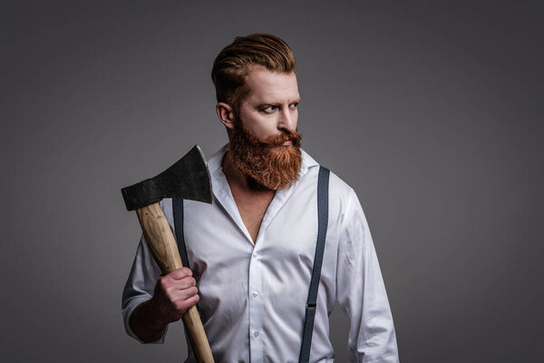 Vintage brutality guy with axe in retro suspenders. Brutal man in suspenders isolated on grey. Mature redhead man with hairstyle. Brutal male fashion style. Male brutality. Extreme brutality.