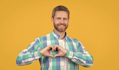 photo of happy man showing love heart. caucasian man gesturing love heart. bearded caucasian man in shirt show love heart isolated on yellow background. caucasian casual man with love heart in studio.